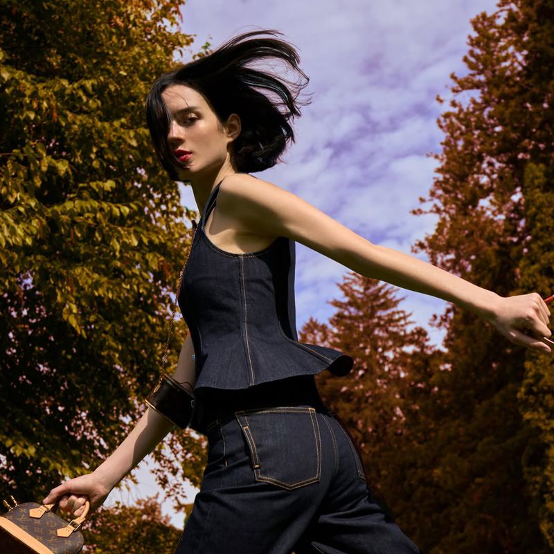 Model Rita Santos wearing a dark denim sleeveless peplum top and matching jeans holding a monogram mini handbag outdoors.