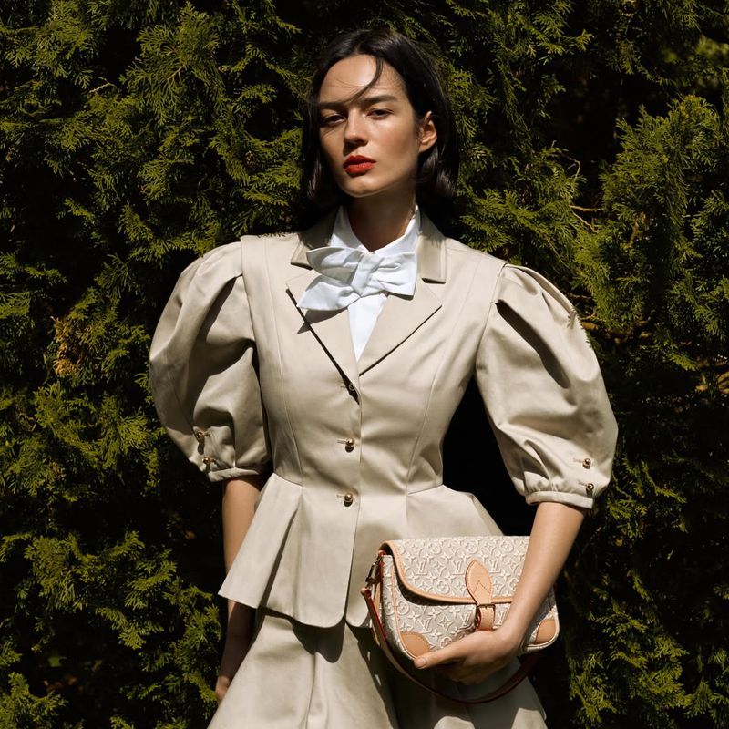 Model wearing a beige puff-sleeve jacket holding a monogram handbag in front of dense green trees