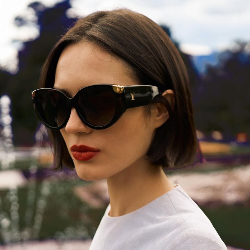 Model with a sleek dark bob wearing oversized black cat-eye sunglasses with gold LV detailing and red lipstick in an outdoor park.