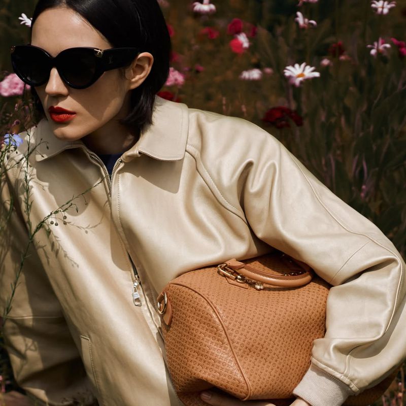 Model Rita Santos wearing a cream leather jacket, oversized black sunglasses, and red lipstick while holding a tan leather handbag.