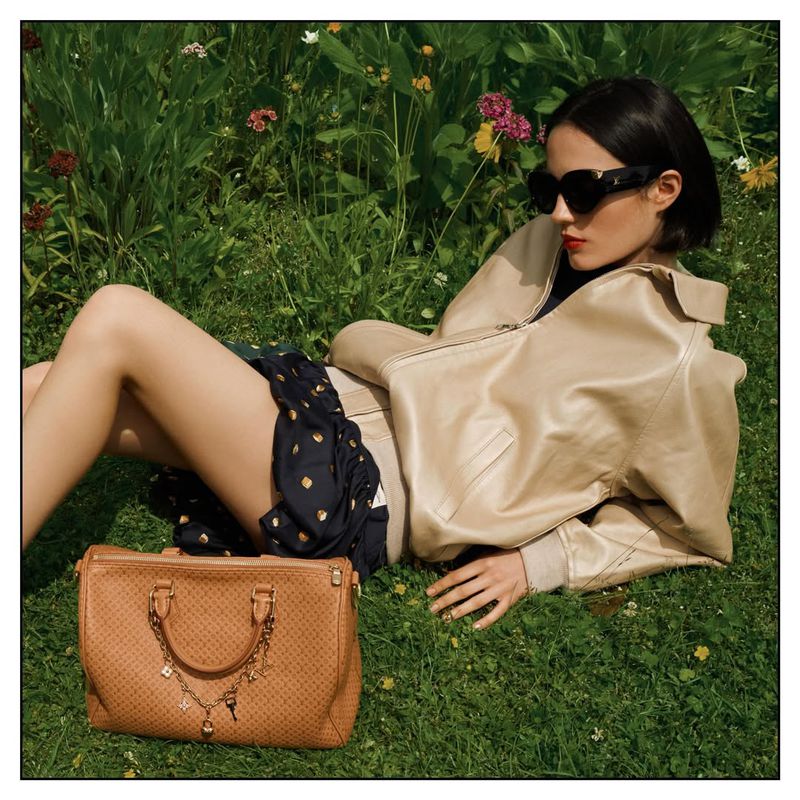 Model Rita Santos in a beige jacket and black sunglasses reclines on a grassy lawn next to a tan leather handbag.