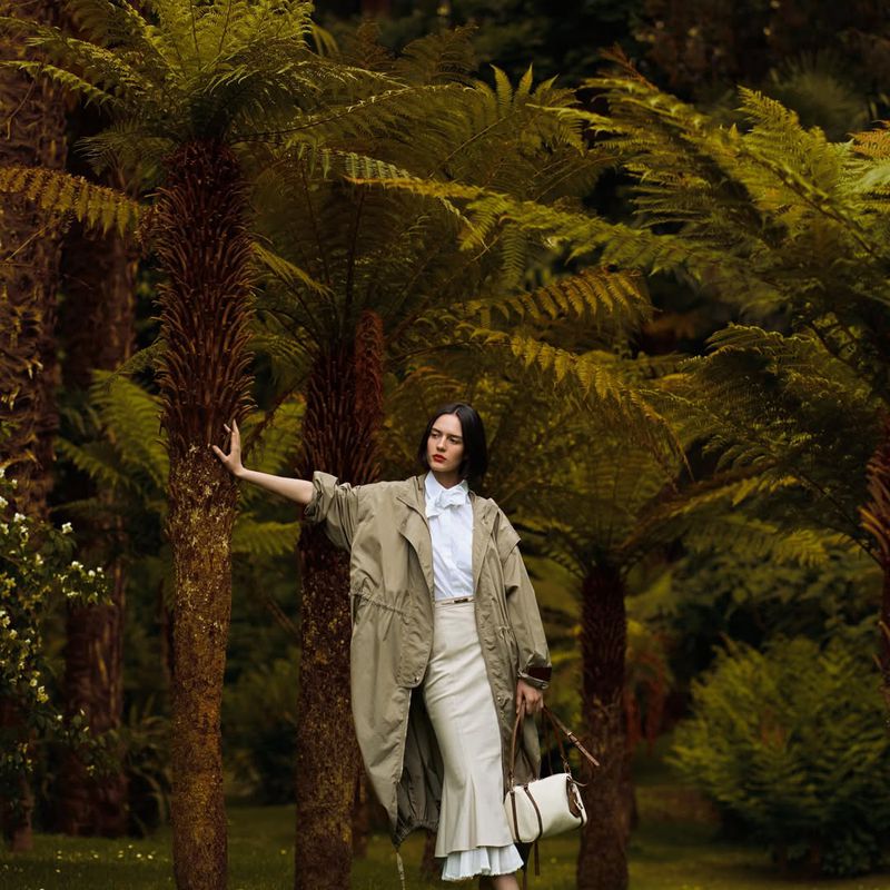 Model Rita Santos in a khaki coat and cream ruffled midi skirt touching a tree trunk amidst lush tree ferns in a garden.