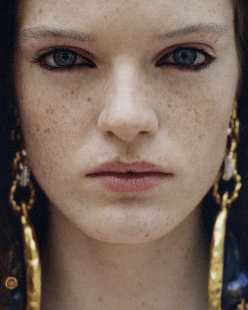 Extreme close-up portrait of a blue-eyed woman with heavy freckles and dark eyeliner wearing large gold chain earrings.