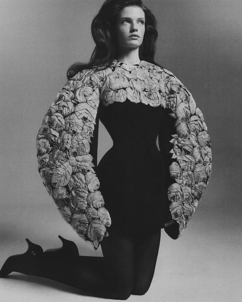 Woman in black dress and floral-embellished bolero kneeling in photo studio wearing high heels, showcasing a fashion-forward editorial look.