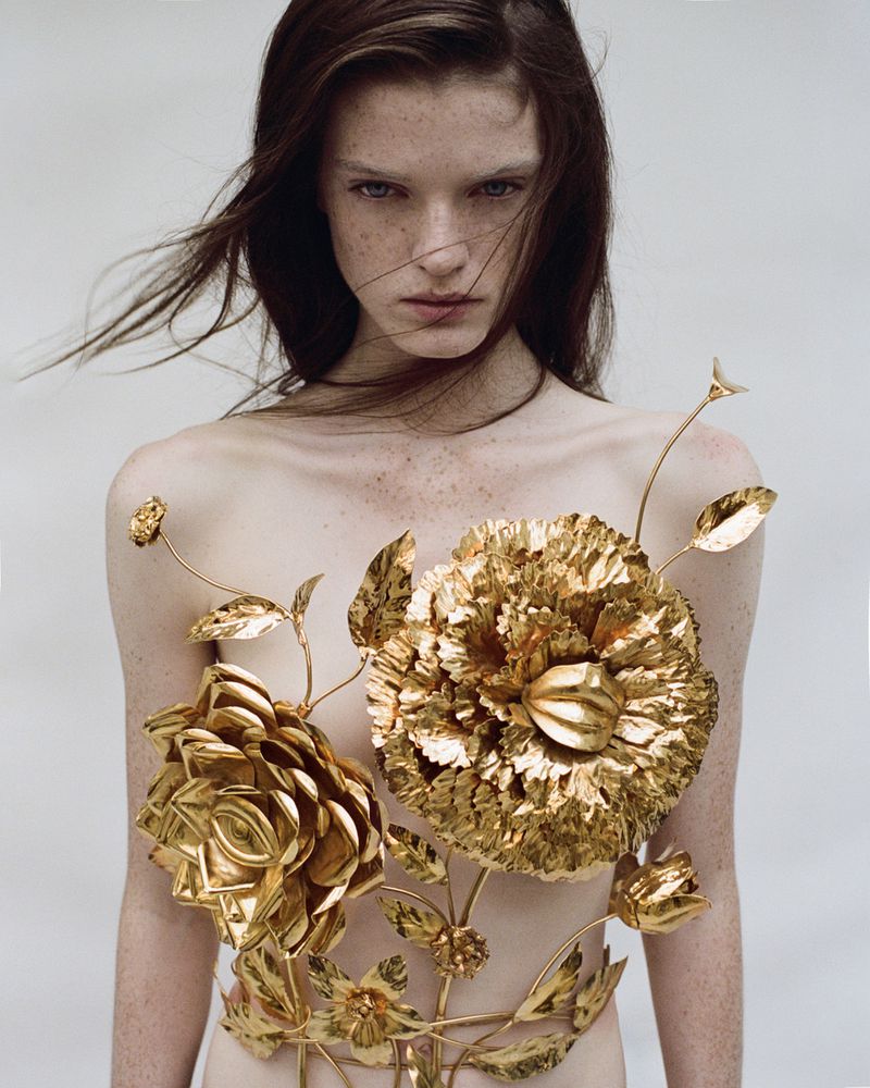 Fashionable portrait of an adult female with fair skin and freckles, posing with golden floral embellishments on bare skin against white background.