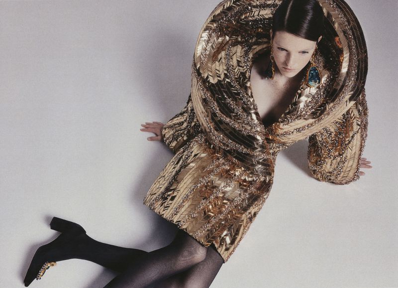 Fashion shot of a brunette adult female wearing a gold sequin dress, black stockings and black heels, posing against a neutral background.