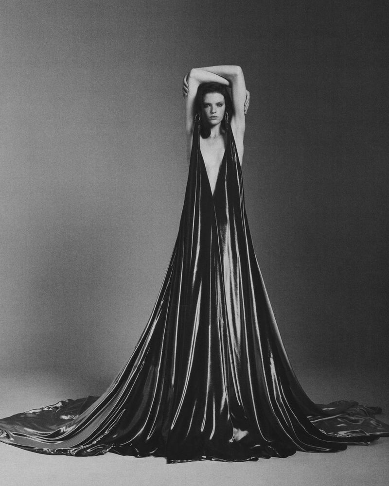 A full-length studio shot of a brunette woman in a flowing black dress, arms raised behind her head, in a monochromatic setting.