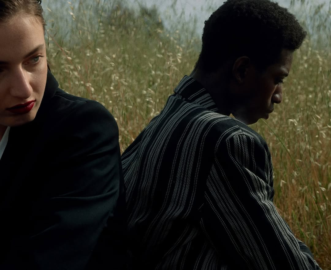 Close-up of a white woman and a black man sitting back to back outdoors amongst tall dry grass in a field.