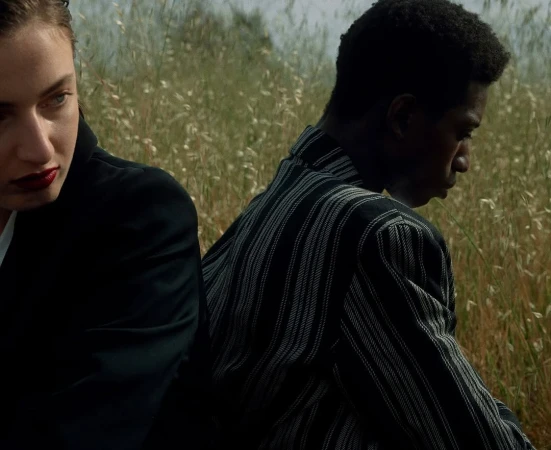 Close-up of a white woman and a black man sitting back to back outdoors amongst tall dry grass in a field.
