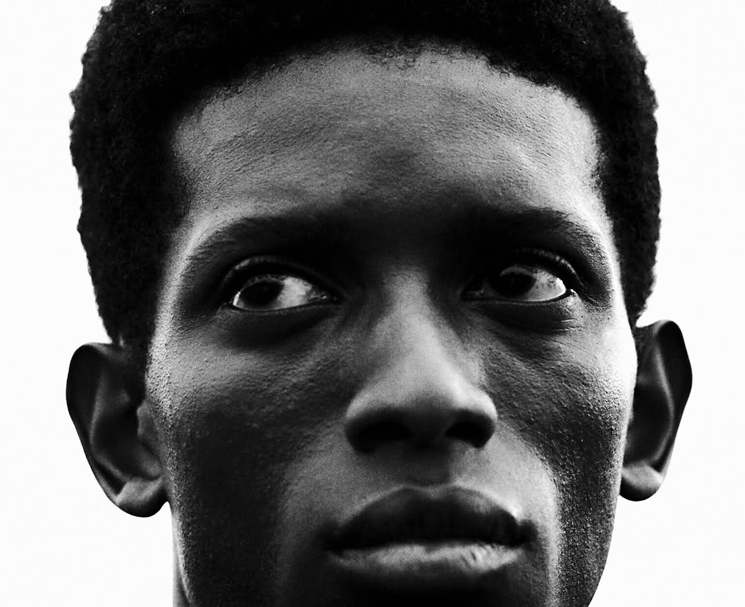 Close-up portrait of a young adult male of African descent with short curly hair and a serious expression against a bright background.