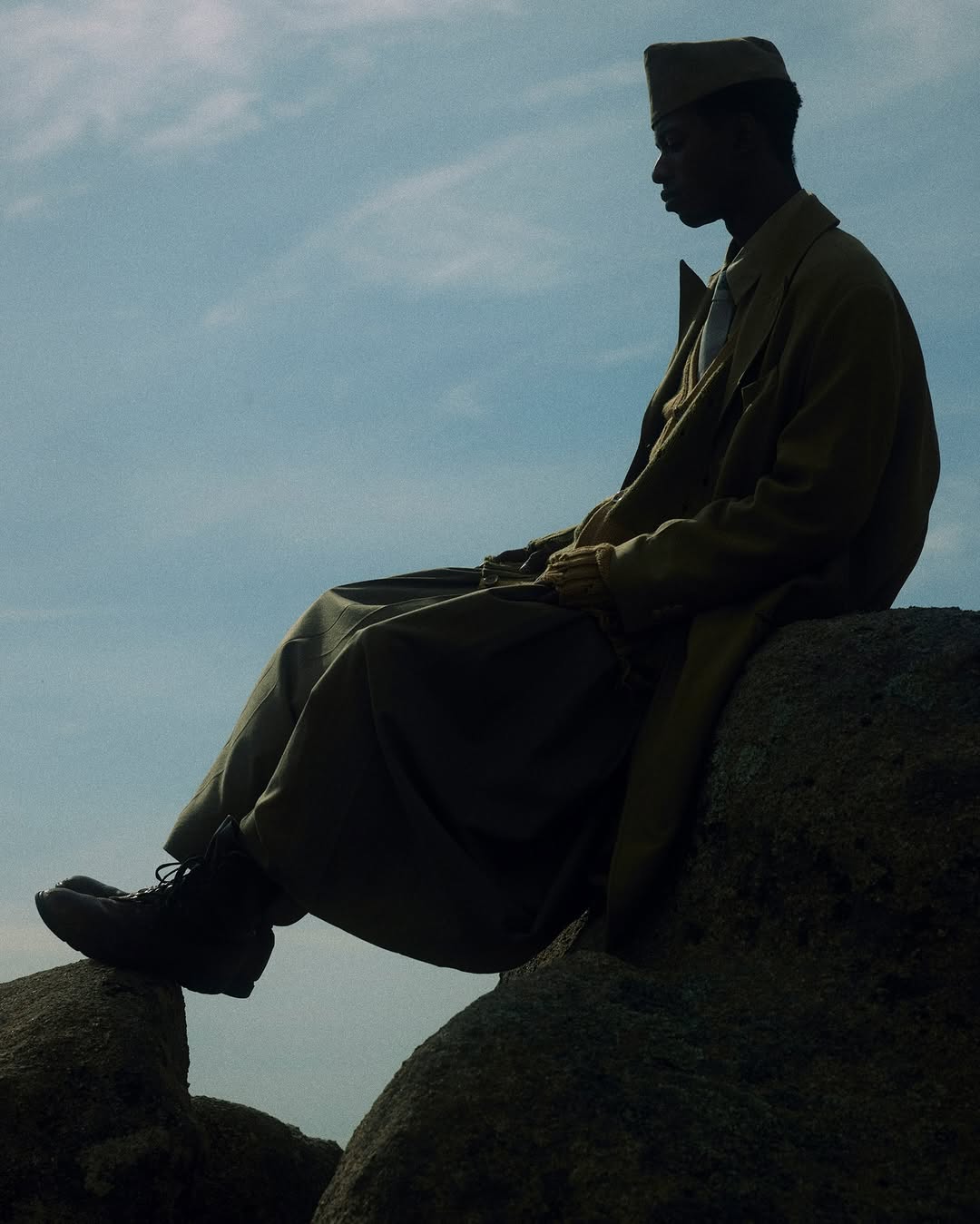 Adult male in an overcoat and cap sits atop large rocks with a partly cloudy sky in the background, creating a serene and contemplative scene.