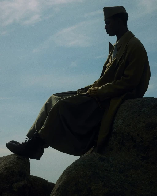 Adult male in an overcoat and cap sits atop large rocks with a partly cloudy sky in the background, creating a serene and contemplative scene.