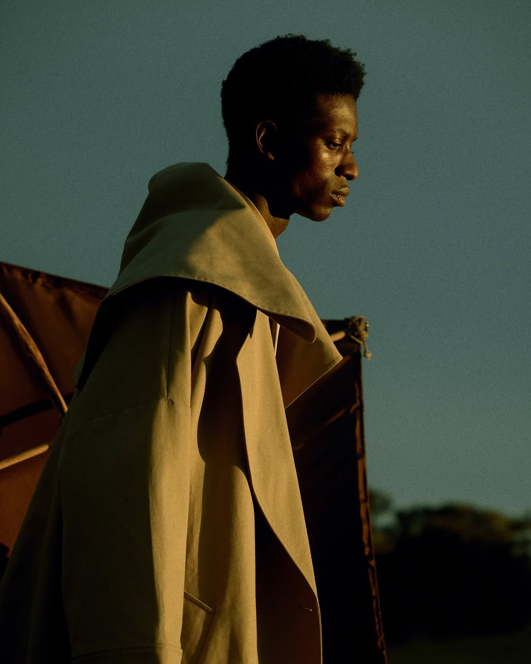 Stylish dark-skinned man in beige trench coat stands outside at dusk with an ambiguous expression. Fashion editorial photography.