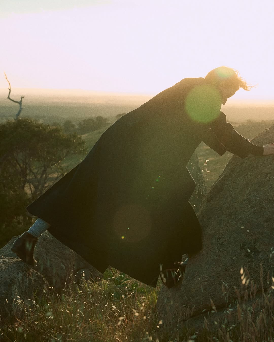 Adult woman in a long dark coat climbing a rocky hill against a blurred background of trees and a light sky during daytime.