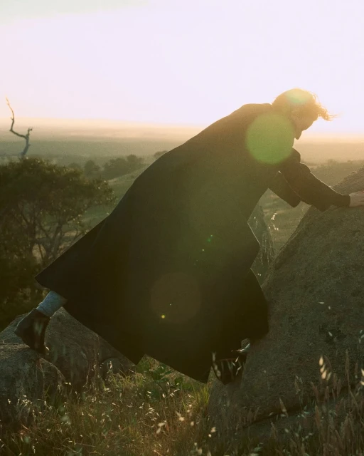 Adult woman in a long dark coat climbing a rocky hill against a blurred background of trees and a light sky during daytime.