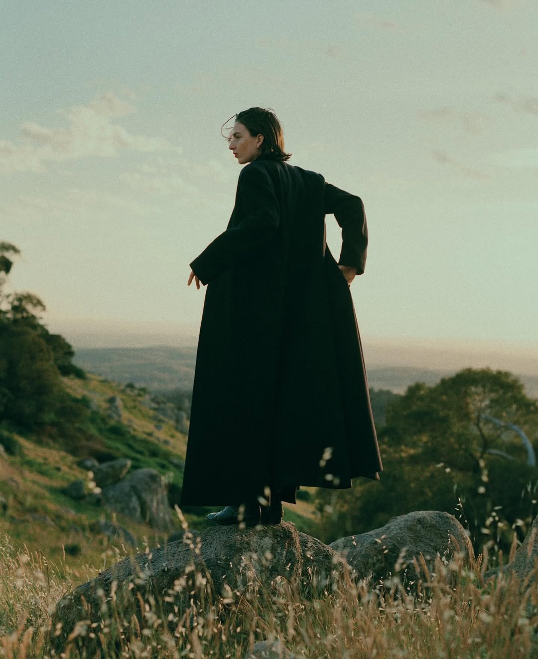 Model standing on a rocky outcrop wearing a long dark coat against a distant landscape background