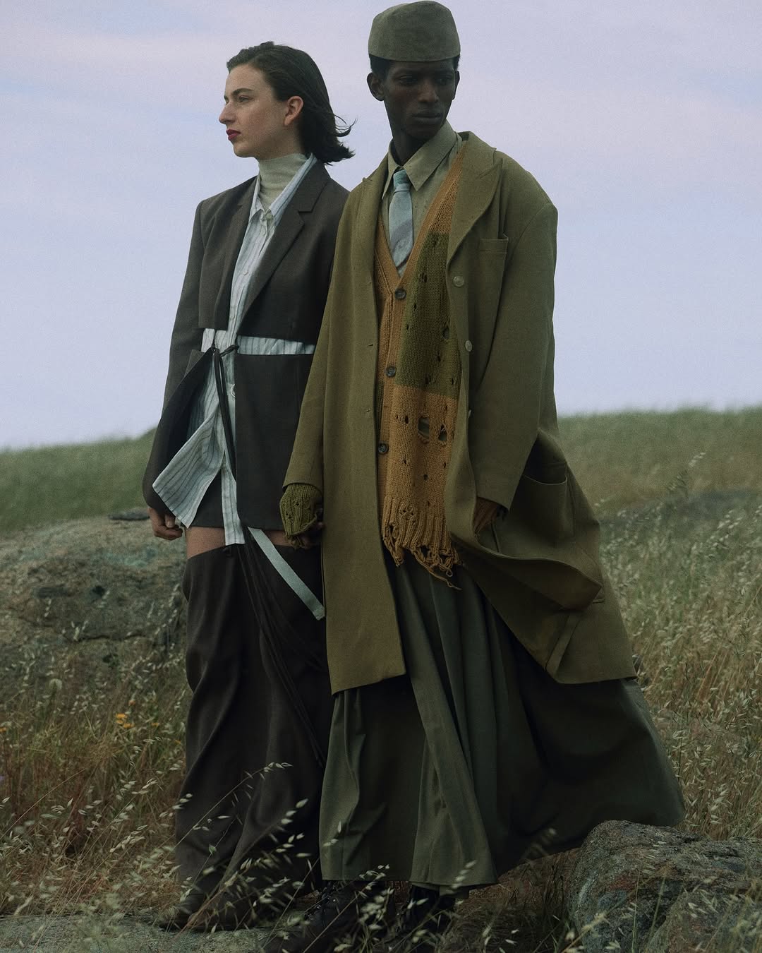 Two fashion models pose in an outdoor landscape. An adult male model and an adult female model stand in a grassy field.