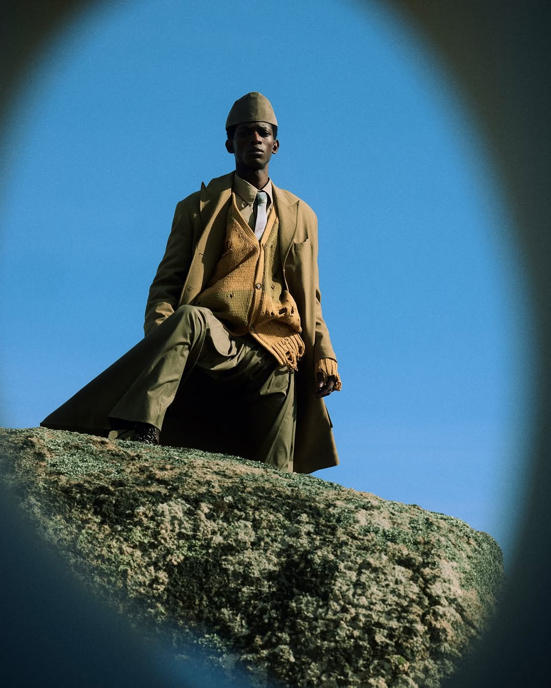 A dark-skinned man sits on a grassy, rocky hill against a clear blue sky wearing a brown coat, knit sweater, tan hat, and khaki trousers.