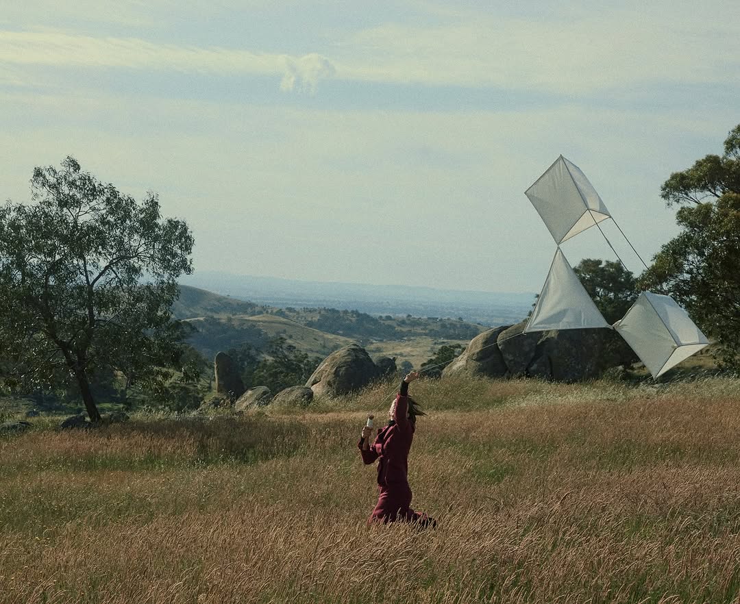 Woman flying a geometric kite in a grassy field with hills and trees in the background, creating a serene outdoor scene.