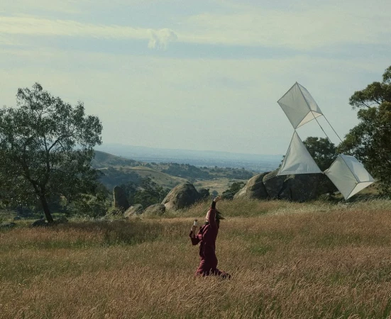 Woman flying a geometric kite in a grassy field with hills and trees in the background, creating a serene outdoor scene.