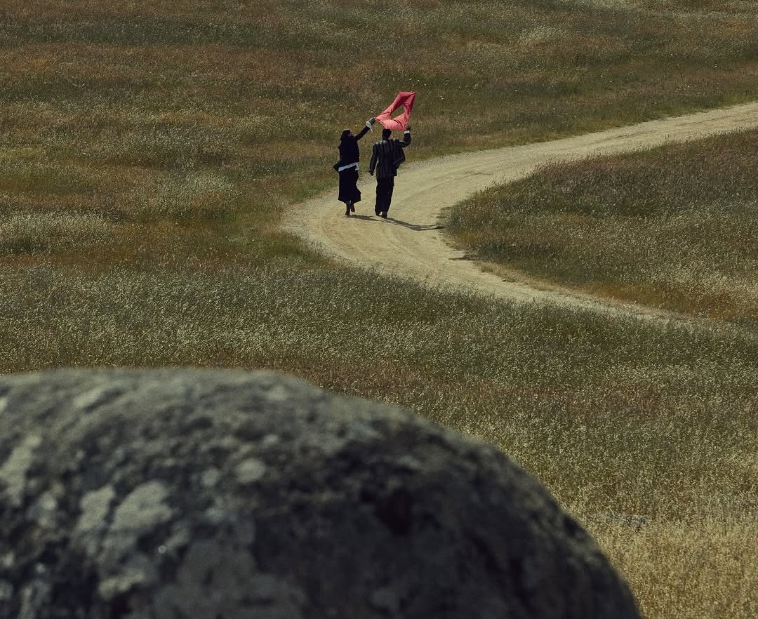 Two adults walk on a dirt path through tall grass, carrying a red object, with a large rock in the foreground.
