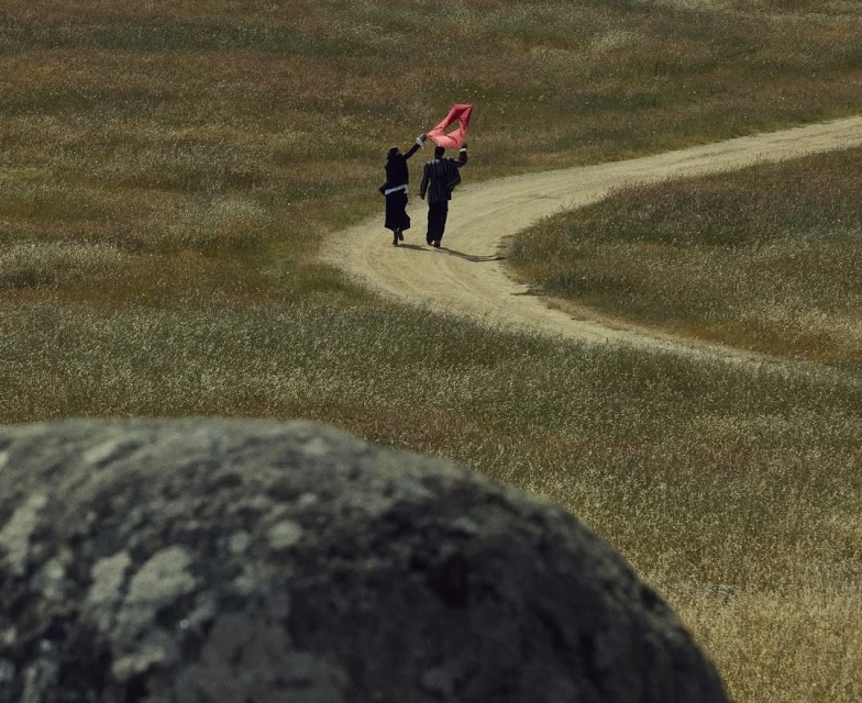 Two adults walk on a dirt path through tall grass, carrying a red object, with a large rock in the foreground.