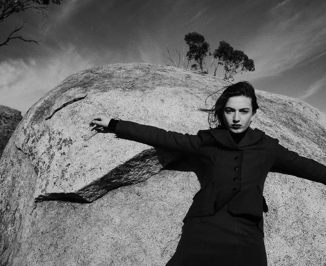 Adult female in dark dress and jacket stands with arms outstretched against a large boulder under a partly cloudy sky in black and white.