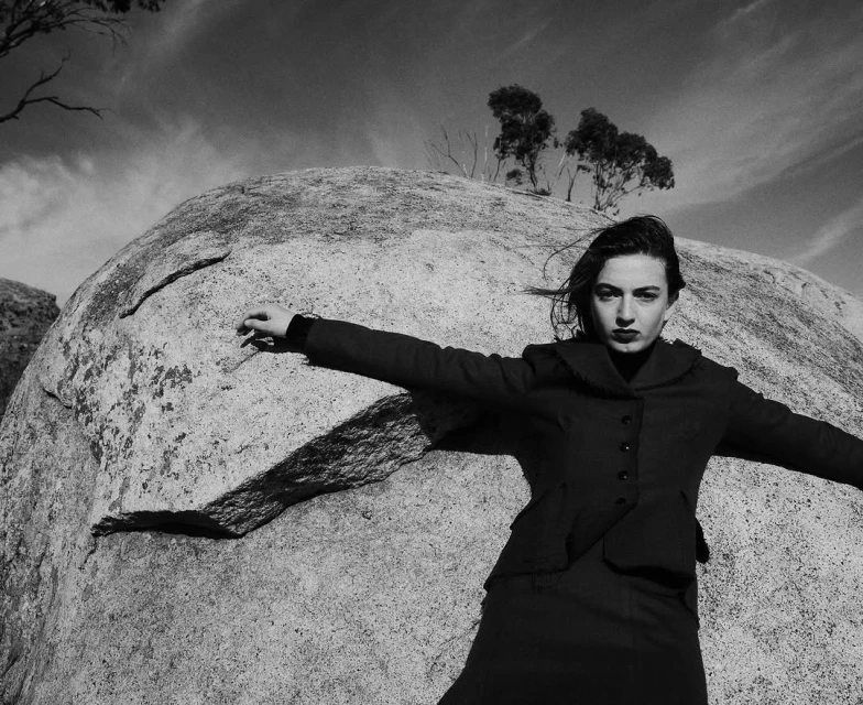 Adult female in dark dress and jacket stands with arms outstretched against a large boulder under a partly cloudy sky in black and white.