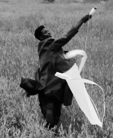 An African American man runs through a grassy field, holding a white kite aloft on a sunny day, in a black and white photograph.