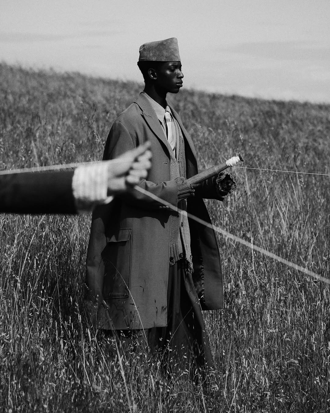 Monochrome image of an African male in a suit and hat holding a fishing rod in a grassy field under a cloudy sky.
