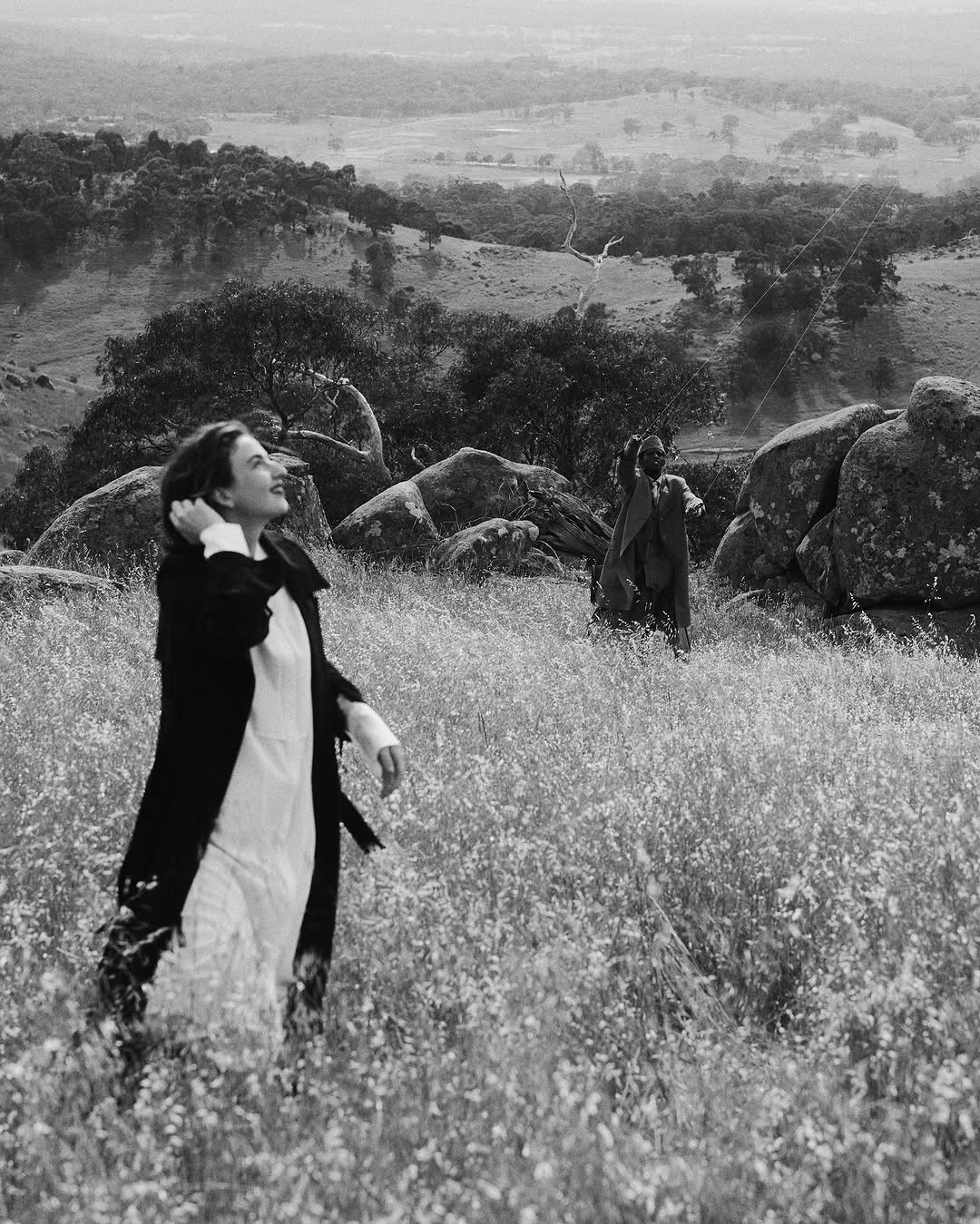 A woman in a white dress and dark coat stands in a field of flowers, with a second woman on a hill in a rural landscape.