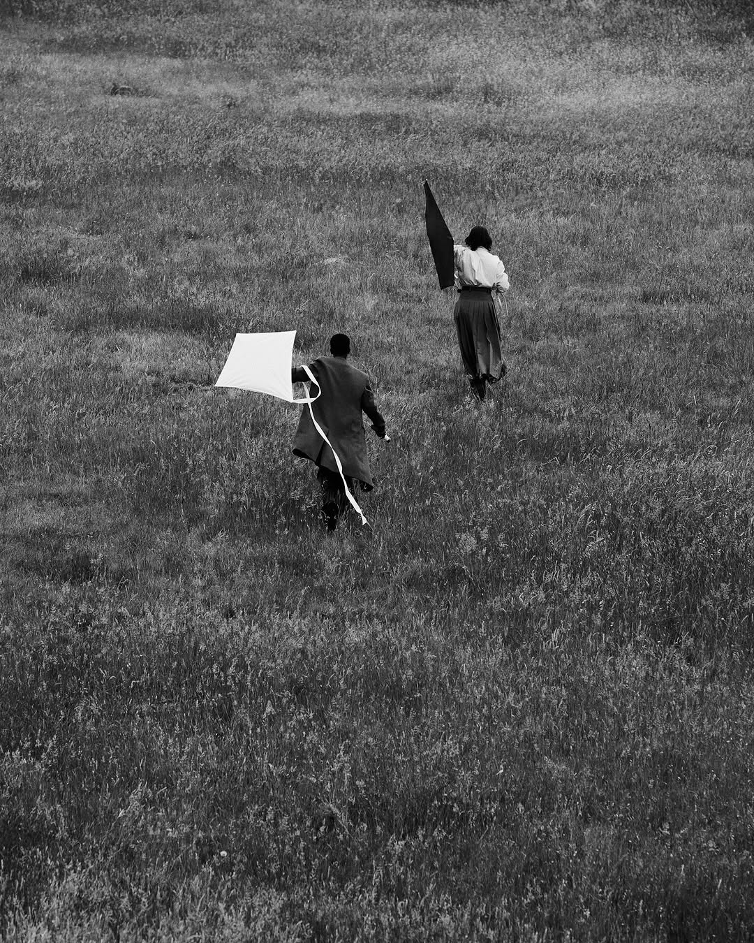 Black and white photo of two people running in a grassy field, flying kites in an open meadow. One person wears a dark coat.