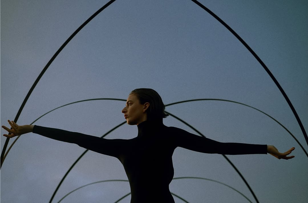Adult woman in black turtleneck poses with arms outstretched against a background of layered arches, creating a striking visual composition.