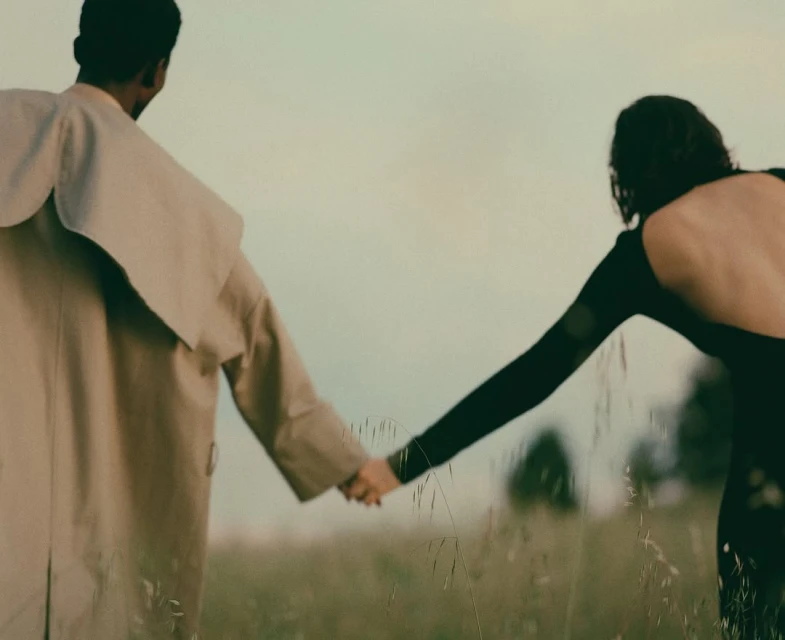 Back view of couple holding hands in field. The woman wears a black dress with open back and man wears a tan trench coat.