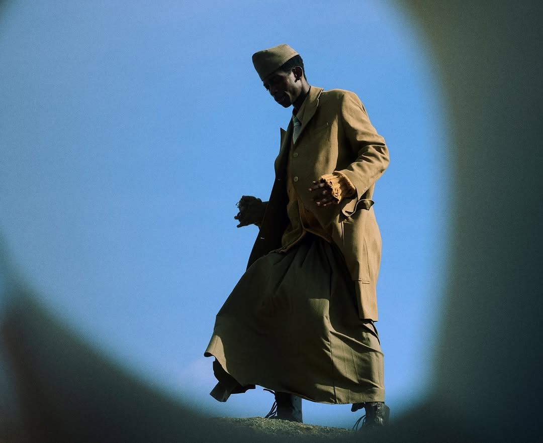 Adult male in a tan suit and hat stands outdoors in front of a clear, light blue sky in a low angle full shot.
