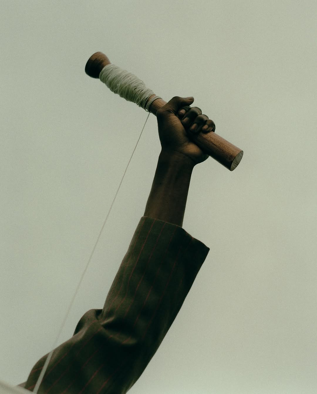 Arm raises a wooden dowel with wound string against pale sky. Dark-skinned adult arm extends, showing a brown jacket sleeve in a low angle shot.