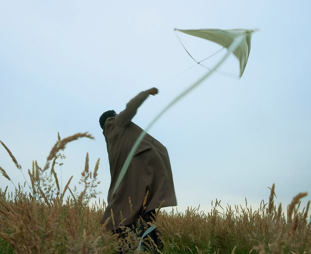 Person in brown coat flying a kite in a field of tall grass under a cloudy sky, creating a serene and nostalgic outdoor scene.