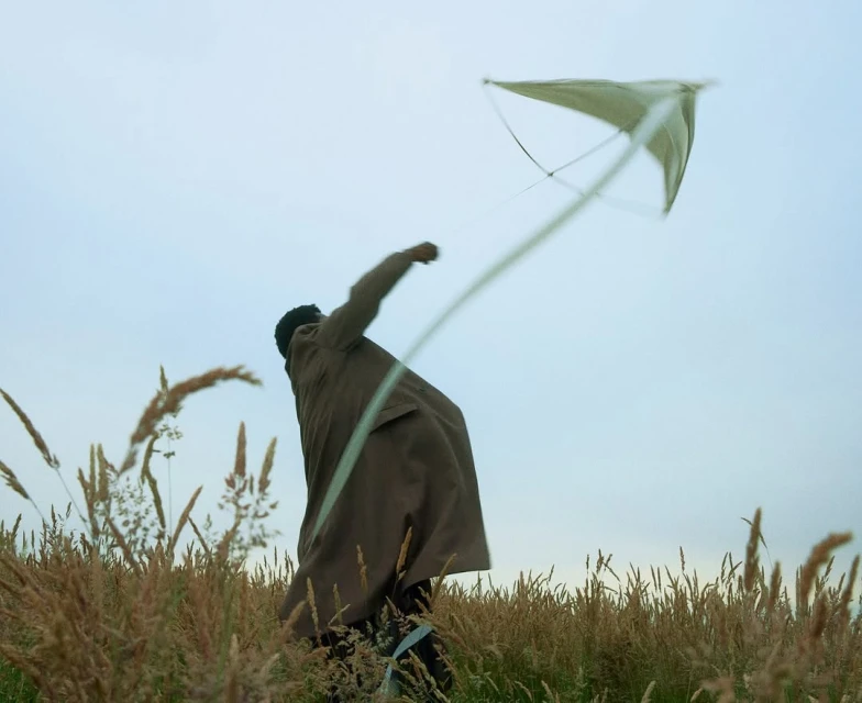 Person in brown coat flying a kite in a field of tall grass under a cloudy sky, creating a serene and nostalgic outdoor scene.