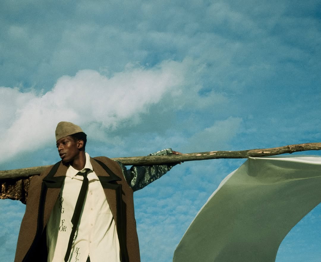 Dark-skinned male model in a tan coat and hat stands with a wooden frame against a sky backdrop, fashion-forward and visually striking.