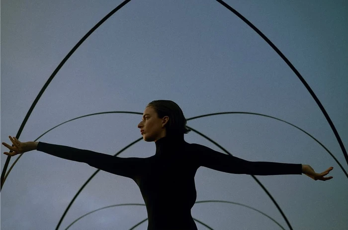 Woman in a black turtleneck posing with outstretched arms beneath curved metal arches against a dusk sky