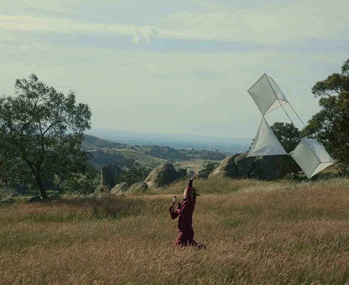 A person running through a grassy field while flying a string of white box kites under a clear sky.