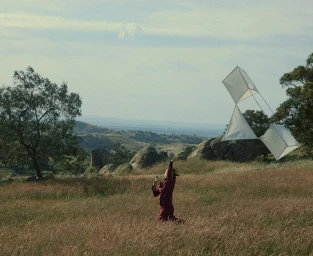 A person running through a grassy field while flying a string of white box kites under a clear sky.