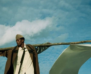 Man wearing a brown coat, patterned shirt, and cap holding a wooden pole against a sky background for Elbazin Magazine