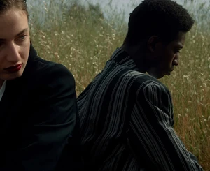 Close-up of a white woman and a black man sitting back to back outdoors amongst tall dry grass in a field.