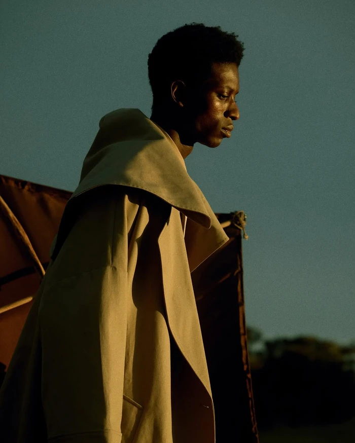 Black man in a camel coat with a large hood standing outdoors against a dark structure during golden hour