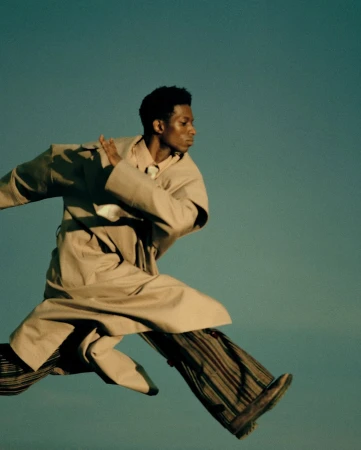 Black man in motion wearing a beige trench coat and patterned trousers against a clear sky background
