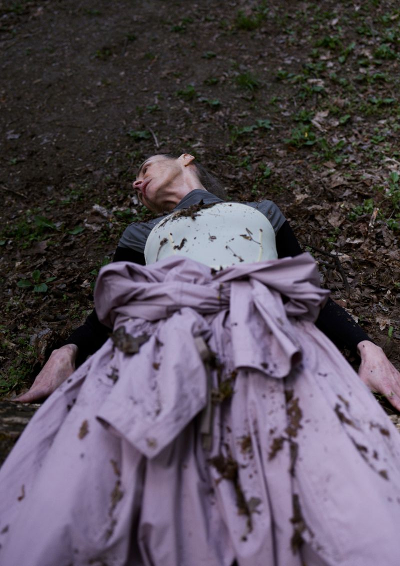 Woman in a pink dress laying on the ground covered in mud, set in an outdoor forest environment with natural lighting.