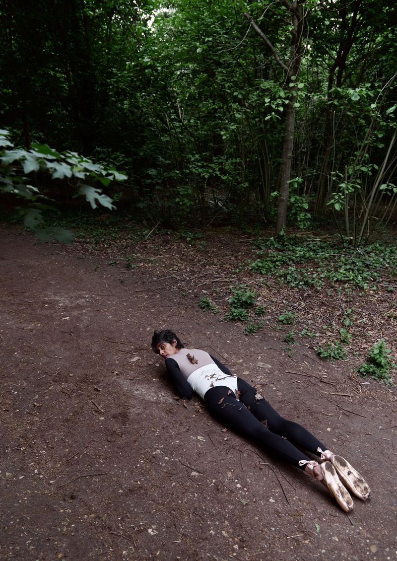 Woman in dancewear lies prone on a dirt path in a green forest setting, wearing black leggings and ballet shoes with ribbons.