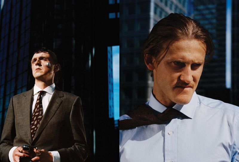 Two men wearing formal business shirts and suits against modern city skyscrapers in bright, hard sunlight.