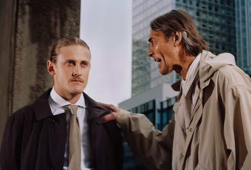 Two men outdoors with an older man gripping the shoulder of a younger man in a dark coat and tie